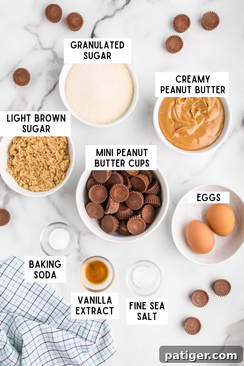 Ingredients on countertop: bowl of granulated sugar, bowl of creamy peanut butter, bowl of light brown sugar, bowl of unwrapped mini reese's peanut butter cups, 2 eggs, baking soda, vanilla, and fine sea salt. Several mini peanut butter cups and a kitchen towel surround the ingredients