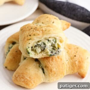 Three spinach artichoke crescent rolls on a plate, one with a bite taken out of it to show the creamy and cheesy spinach dip inside of it.