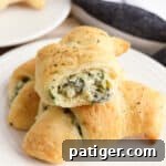 Three spinach artichoke crescent rolls on a plate, one with a bite taken out of it to show the creamy and cheesy spinach dip inside of it.