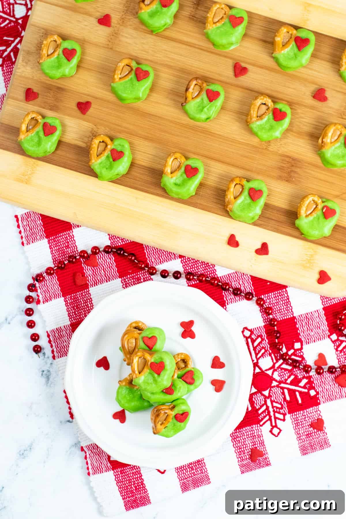 Grinch Pretzel Bites on white plate with red heart sprinkles sprinkled around them and a tray of neatly arranged pretzel bites in background