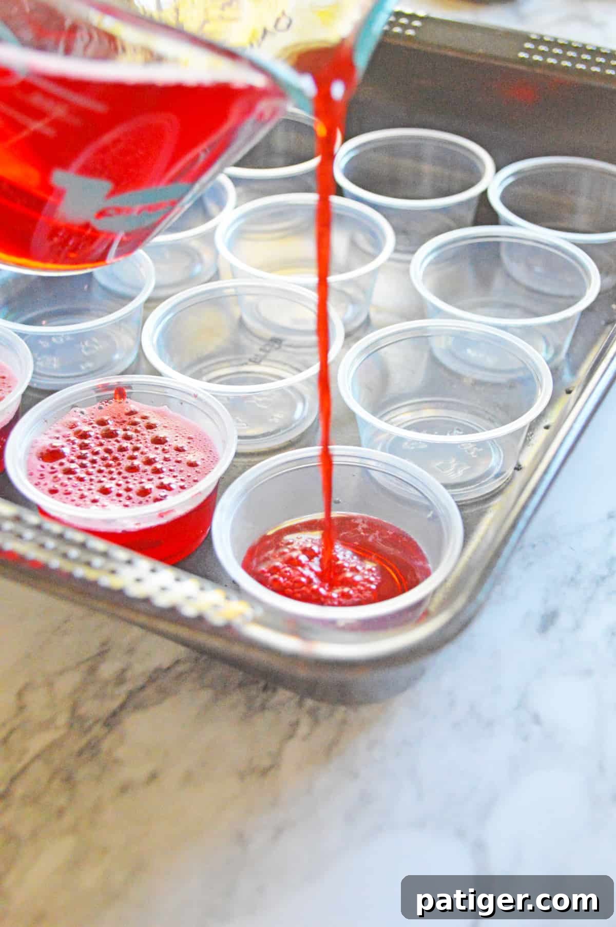 Strawberry jello shots being poured into small plastic shot cups arranged in a baking pan for stability.