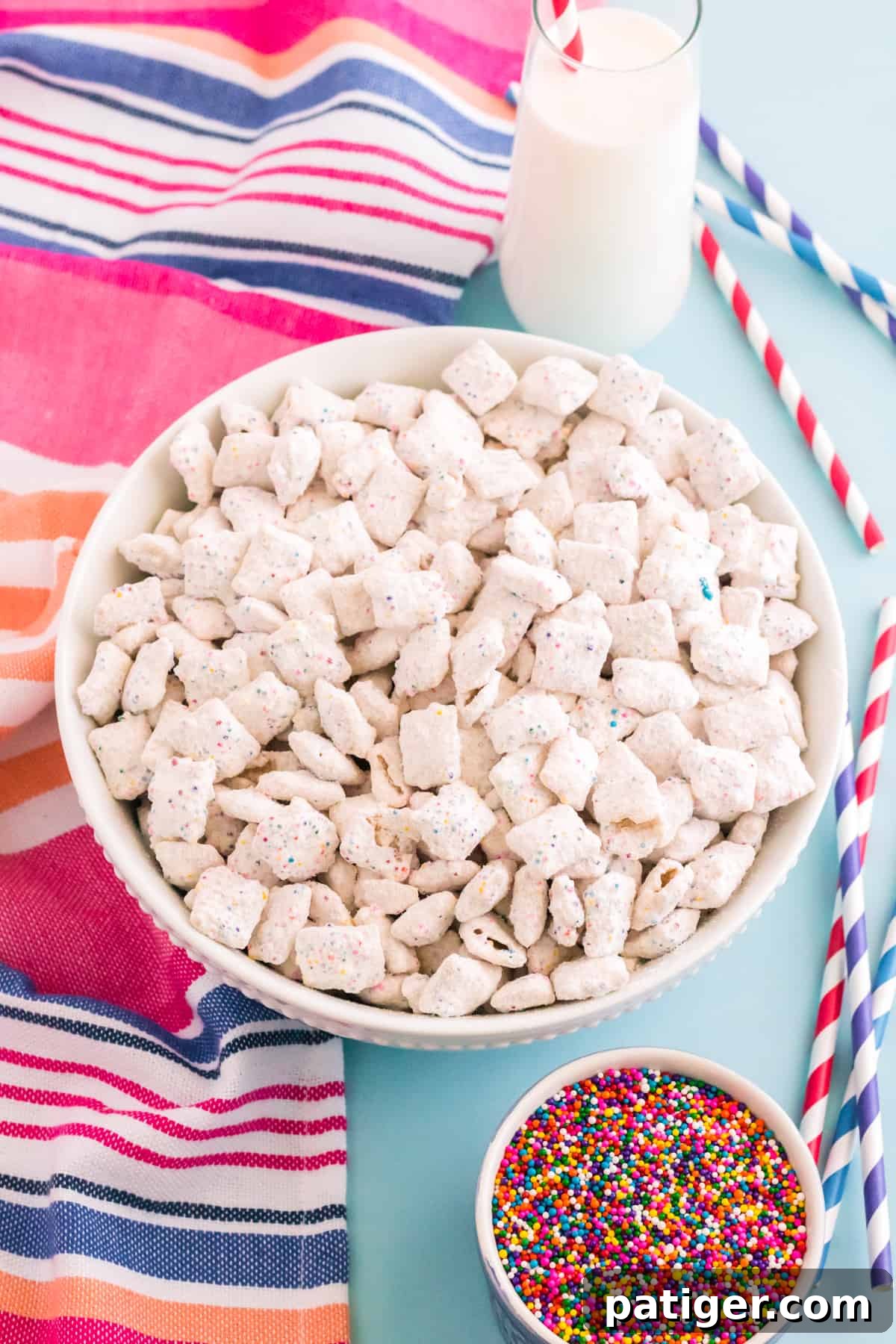 Birthday cake puppy chow in large white bowl with glass of milk, rainbow sprinkles, and colorful linen and straws surrounding it.