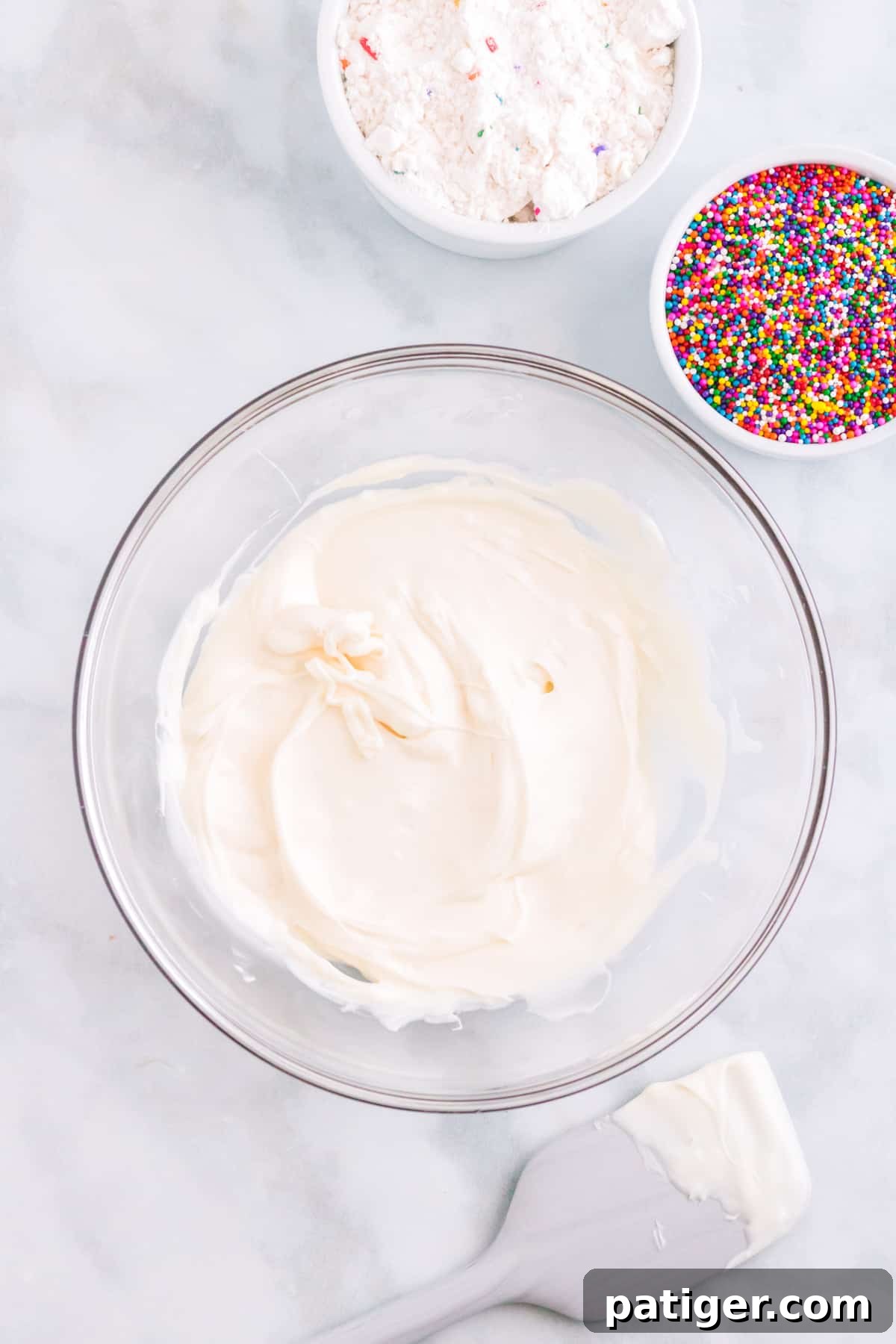 Melted white candy melts in glass bowl with rainbow sprinkles, white chocolate covered spatula, and funfetti cake mix next to it