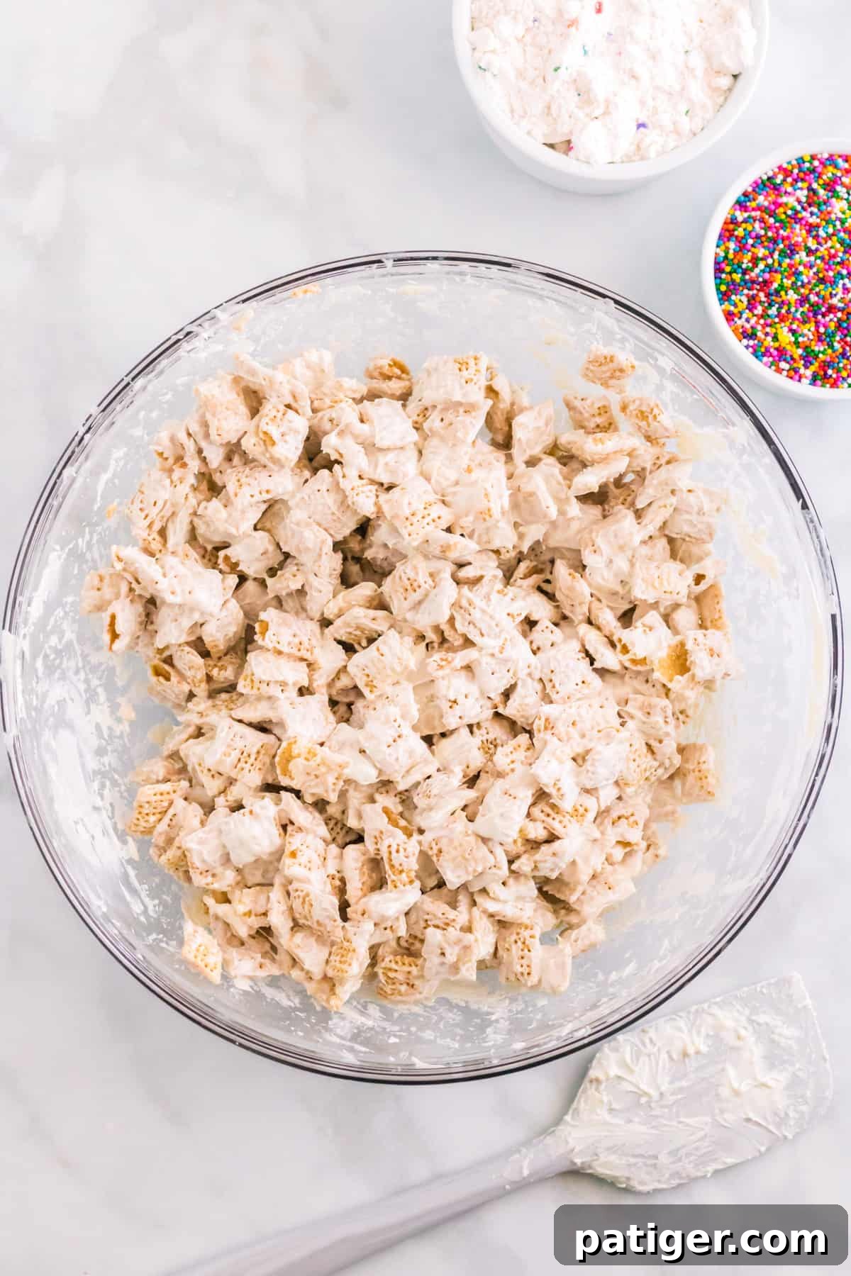 White candy melt coated chex cereal in large glass bowl with rainbow sprinkles, spatula, and funfetti cake mix beside it