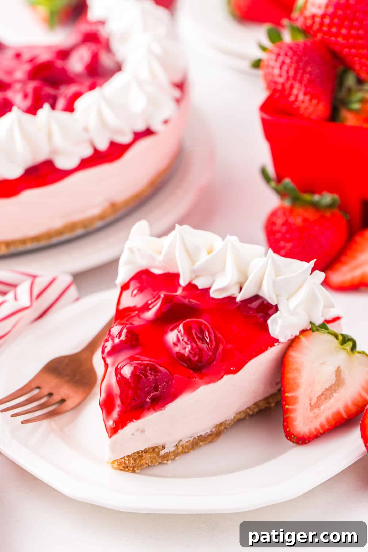 Slice of no bake strawberry cheesecake with strawberry topping and whipped cream piping on edge. Remaining cake and fresh strawberries are in background.