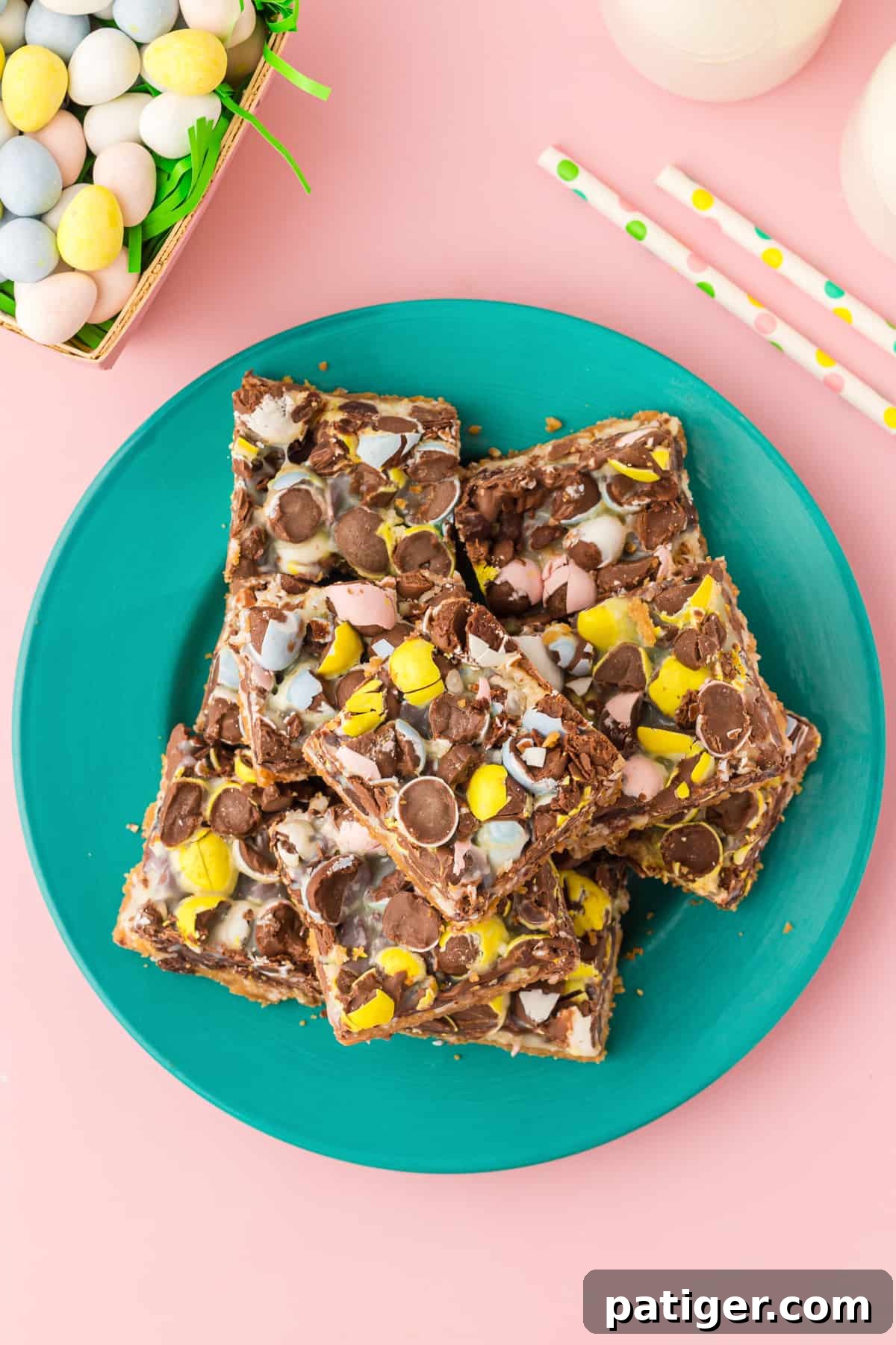 Top-down photo of mini egg cookie bars just into squares and laid on teal plate. Mini chocolate eggs, paper straws, and glasses of milk are in background.
