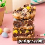 Easter dessert bars with sweetened condensed milk, chocolate chips, coconut, and chocolate eggs stacked on top of one another with top bar missing a bite.