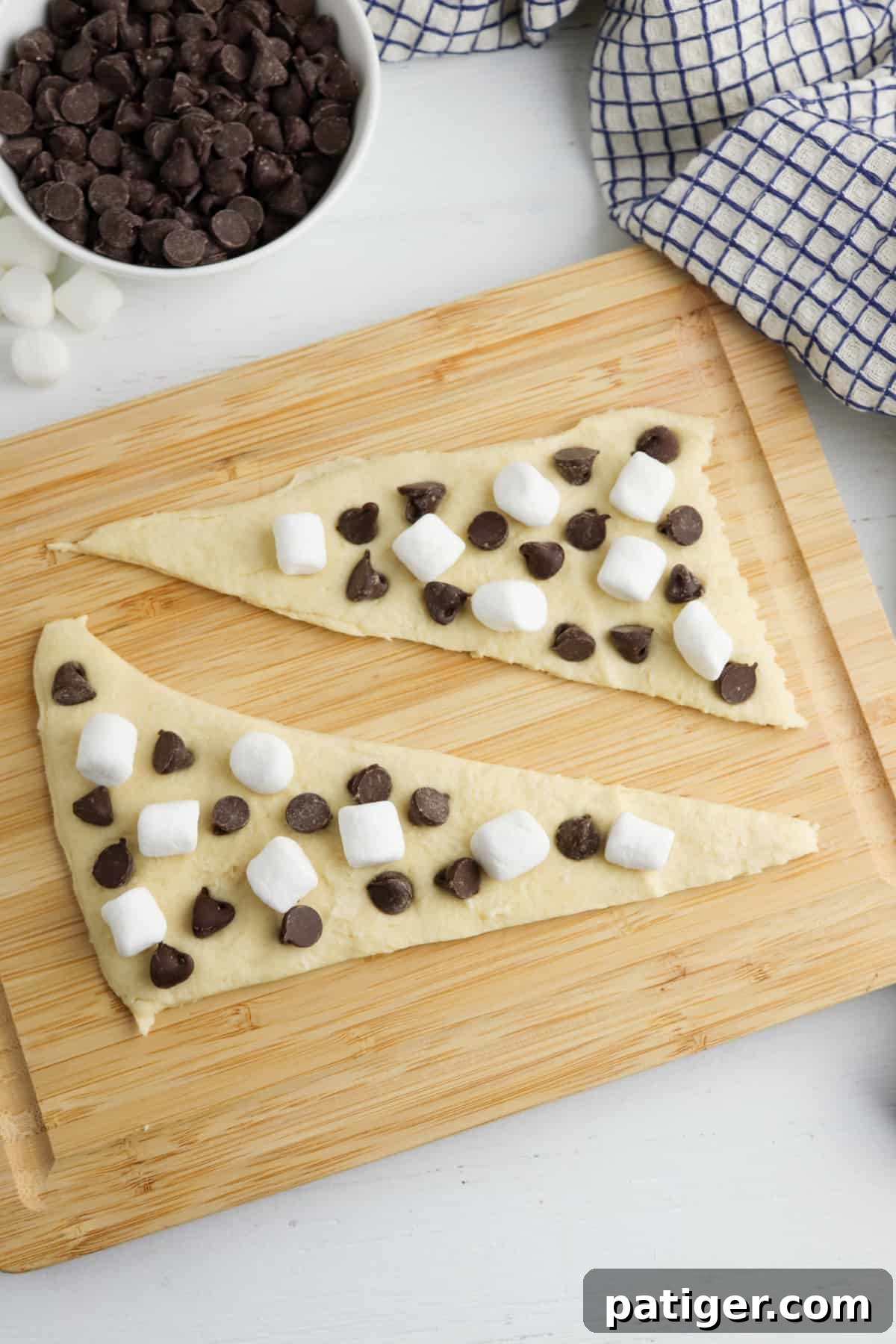 Two triangles of crescent roll dough laid flat on a cutting board, generously topped with mini marshmallows and chocolate chips, ready to be rolled.