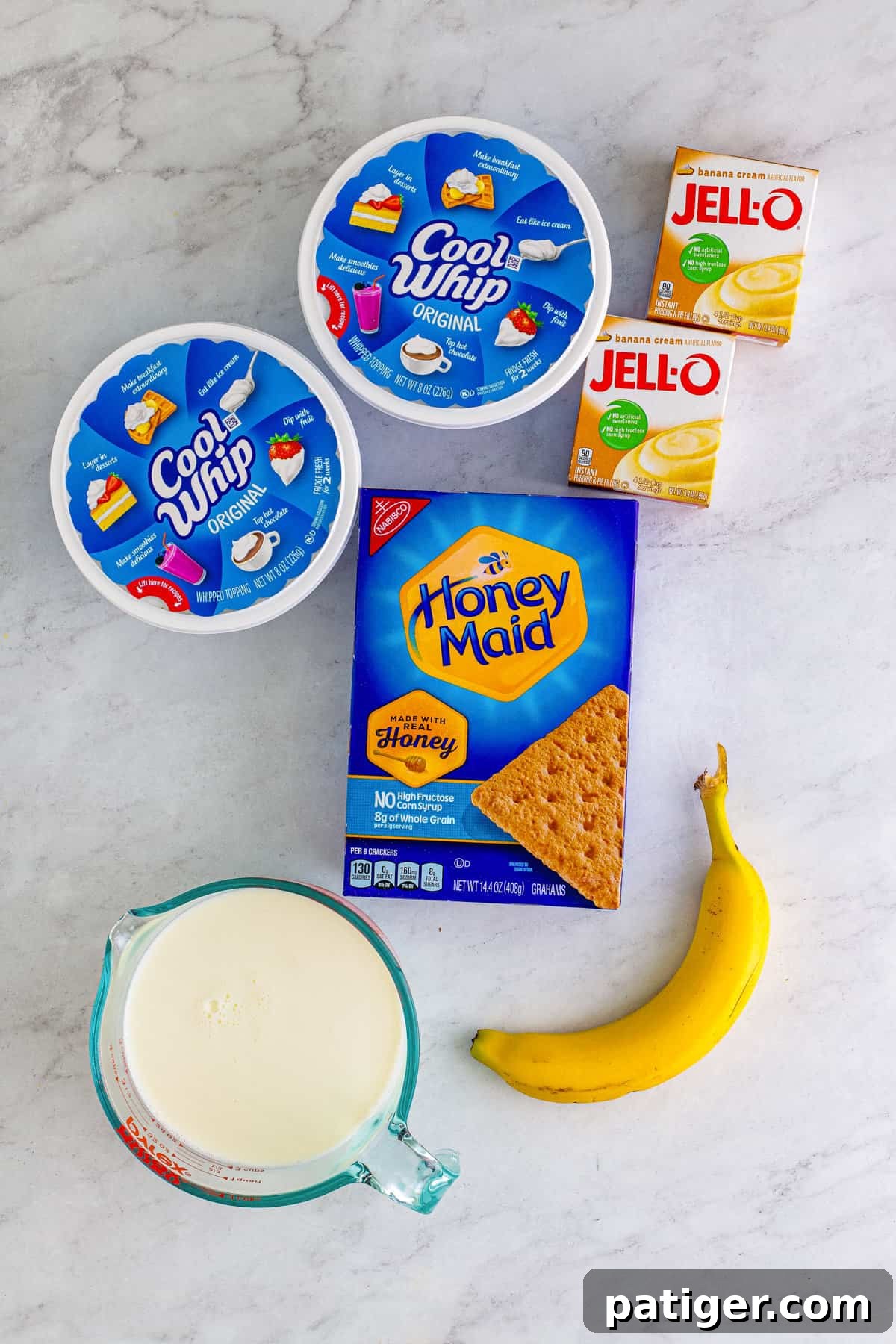 Two containers of Cool Whip, 2 boxes of vanilla instant pudding mix, a box of honey maid graham crackers, a banana, and a measuring cup of milk.