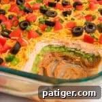 A partially scooped 7 layer dip without cream cheese, highlighting colorful layers of bean dip, sour cream, and guacamole, and garnished with fresh tomatoes, olives, and green onions. The inviting image shows the creamy texture and vibrant appearance.