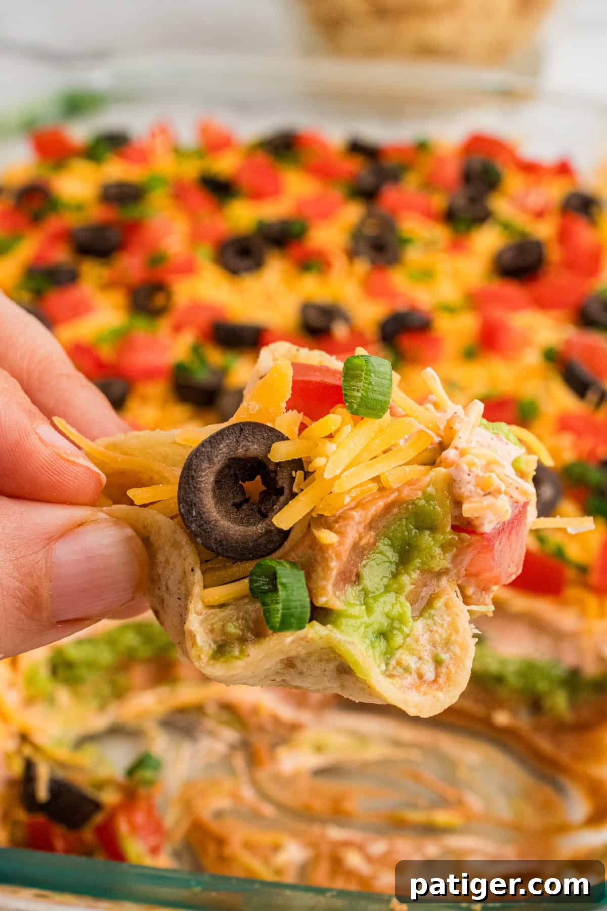 A close-up of a crunchy tortilla chip generously loaded with 7-layer taco dip. The distinct, colorful layers of refried beans, vibrant guacamole, creamy sour cream, shredded cheese, fresh tomatoes, sliced black olives, and green onions are clearly visible and appetizing.