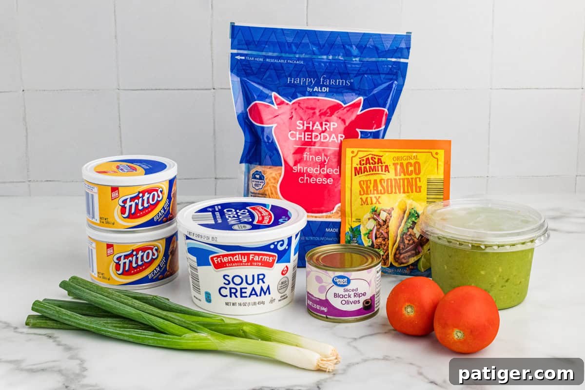 A countertop displays all the fresh and packaged ingredients needed for a 7-layer taco dip. These include two cans of Fritos bean dip, a tub of sour cream, a bag of shredded sharp cheddar cheese, a packet of taco seasoning mix, a container of guacamole, a can of sliced black olives, fresh Roma tomatoes, and a bunch of green onions.