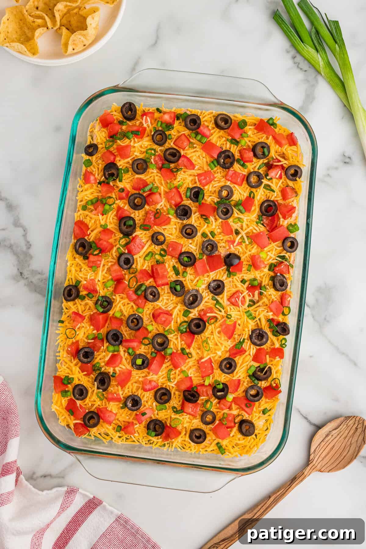 A clear 9x13 glass dish filled with a vibrant 7-layer taco dip, made without cream cheese. The top layers are beautifully adorned with a generous sprinkle of shredded cheddar cheese, fresh diced tomatoes, glossy black olives, and thinly sliced green onions, showcasing its readiness for a party.