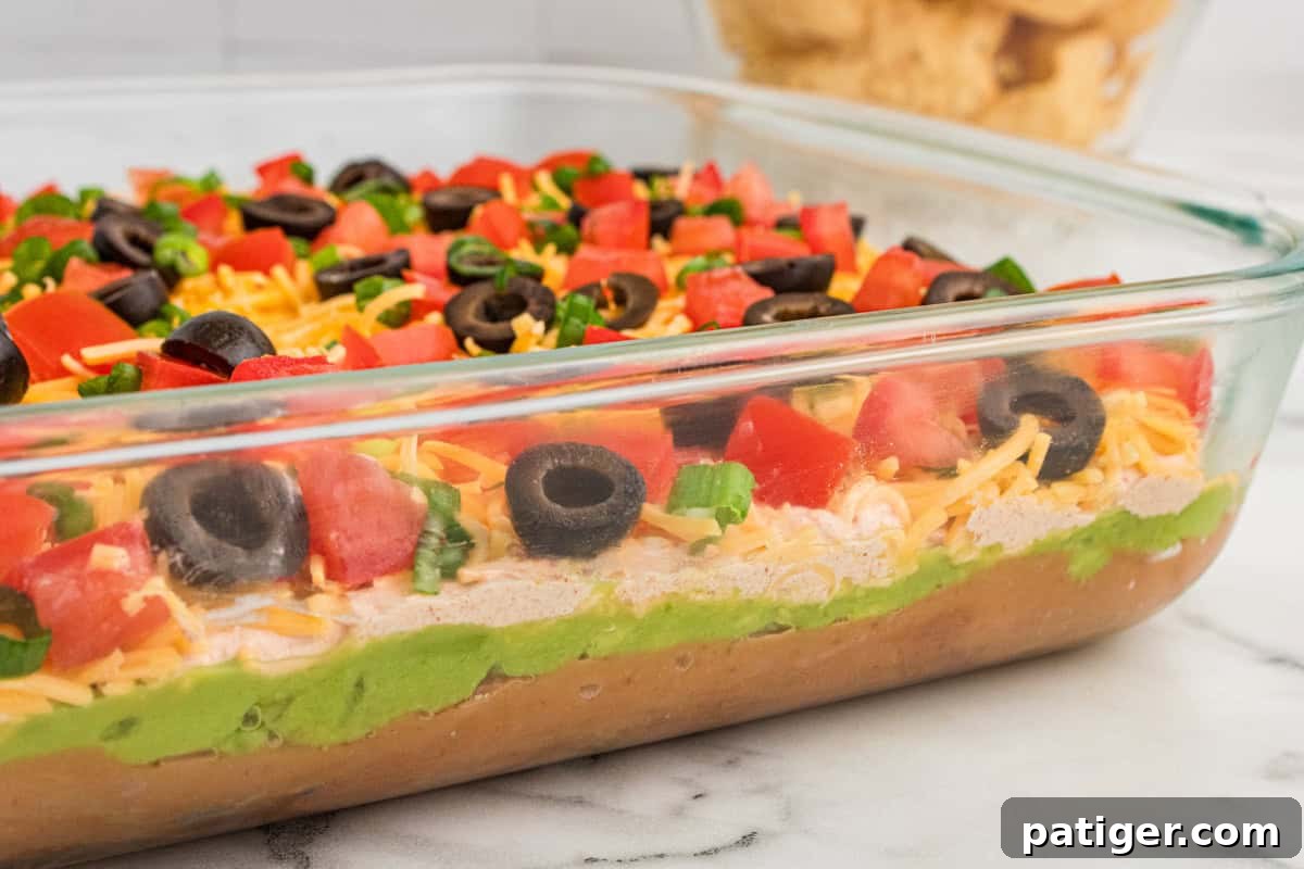 A side view of a 9x13 glass dish filled with a perfectly layered 7-layer dip. Clearly visible are distinct strata of refried beans, vibrant guacamole, taco-seasoned sour cream, a blanket of shredded cheese, and fresh toppings including diced tomatoes, black olives, and green onions, highlighting the artistry of this appetizer.