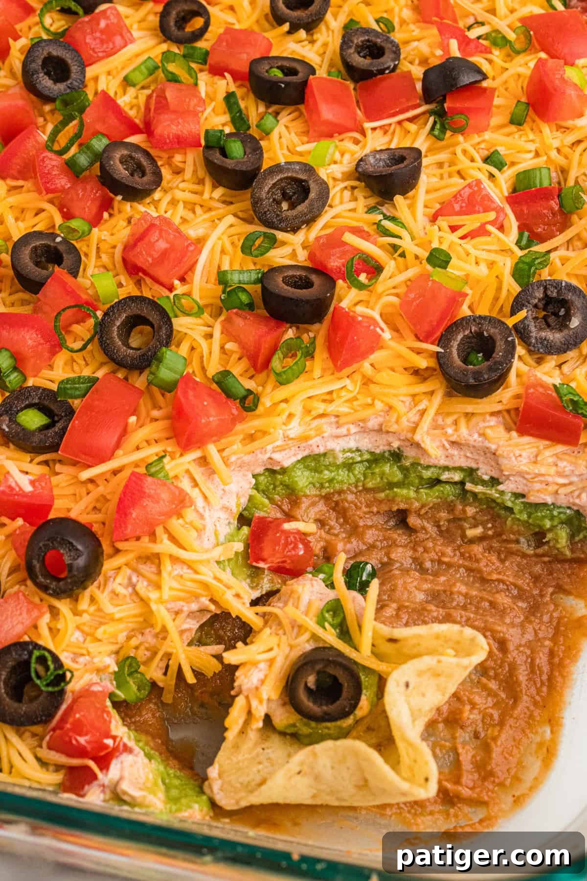 A scoop has been taken from a 7-layer taco dip, clearly revealing the distinct and delicious layers of bean dip, creamy guacamole, and seasoned sour cream. The dip is presented in a glass dish, highlighting its inviting texture and vibrant colors.