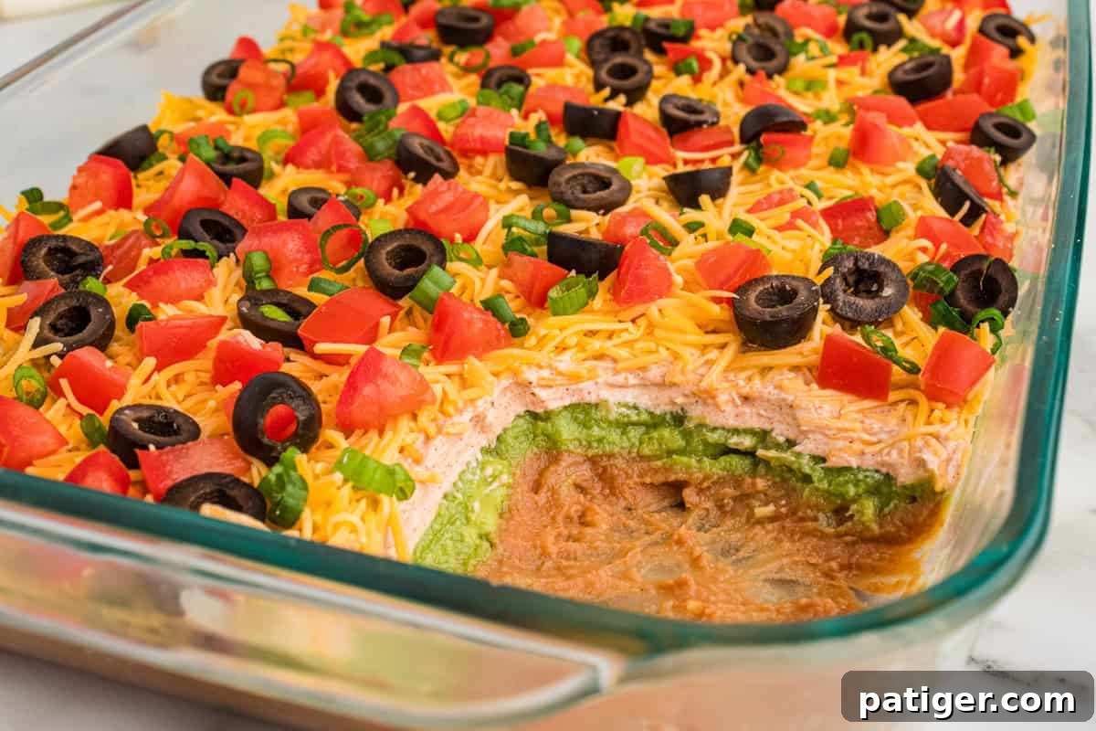 A close-up view of a partially scooped 7-layer dip in a clear glass dish, highlighting the distinct and appetizing layers of refried beans, vibrant guacamole, perfectly seasoned sour cream, shredded cheddar cheese, and a colorful array of diced tomatoes, black olives, and green onions on top.