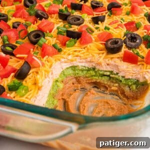 A partially scooped 7 layer dip without cream cheese, highlighting colorful layers of bean dip, sour cream, and guacamole. It is garnished with fresh tomatoes, olives, and green onions, ready to be served.