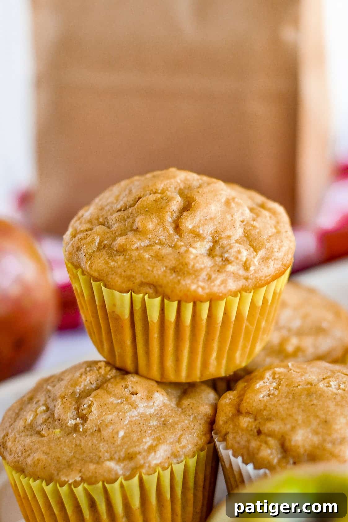 Freshly baked apple oat muffins stacked elegantly in front of a rustic brown paper lunch bag, showcasing their golden tops and inviting texture.
