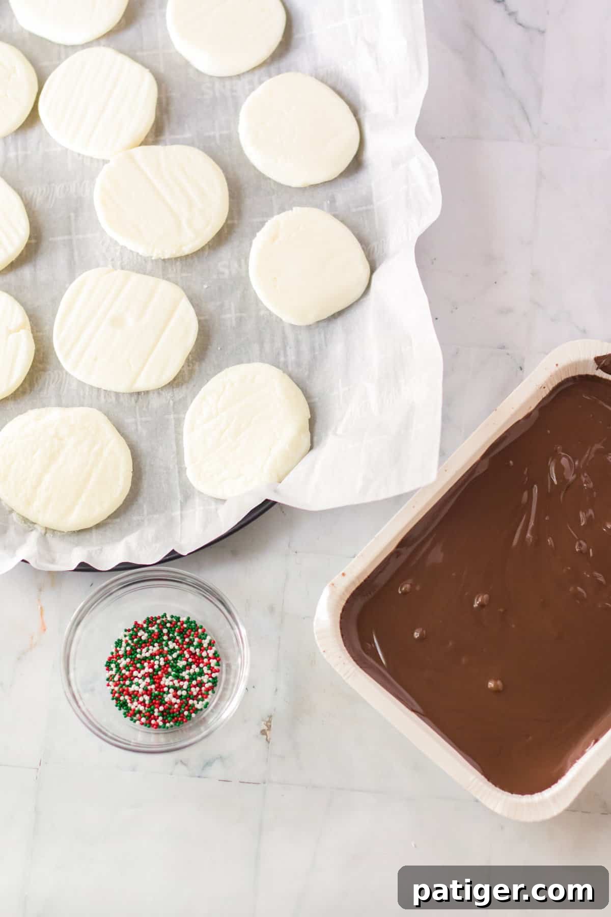 Alongside a bowl of glossy melted almond bark and a scattering of colorful sprinkles, perfectly frozen peppermint discs are laid out, ready for their chocolate coating.