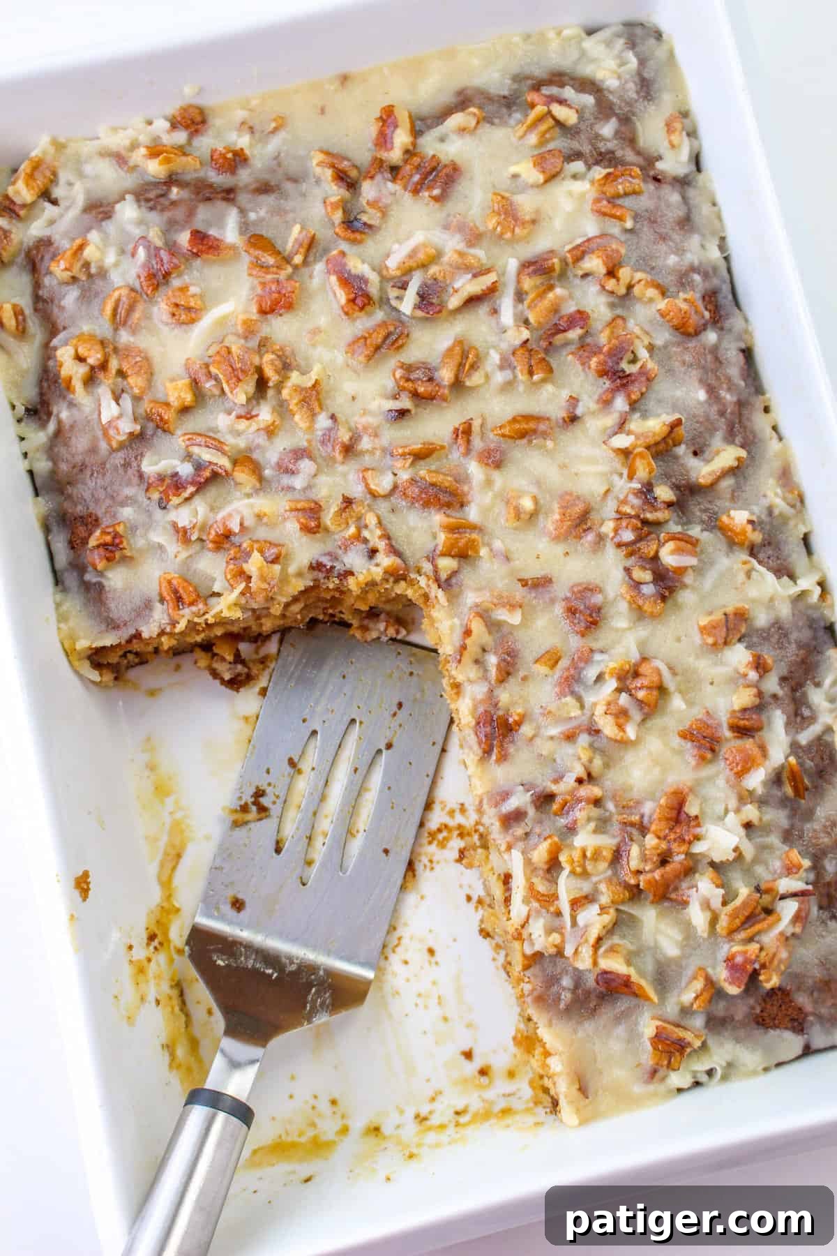 Fruit cocktail cake with coconut pecan frosting in a 9 x 13 baking dish with a few slices removed and spatula resting in dish.