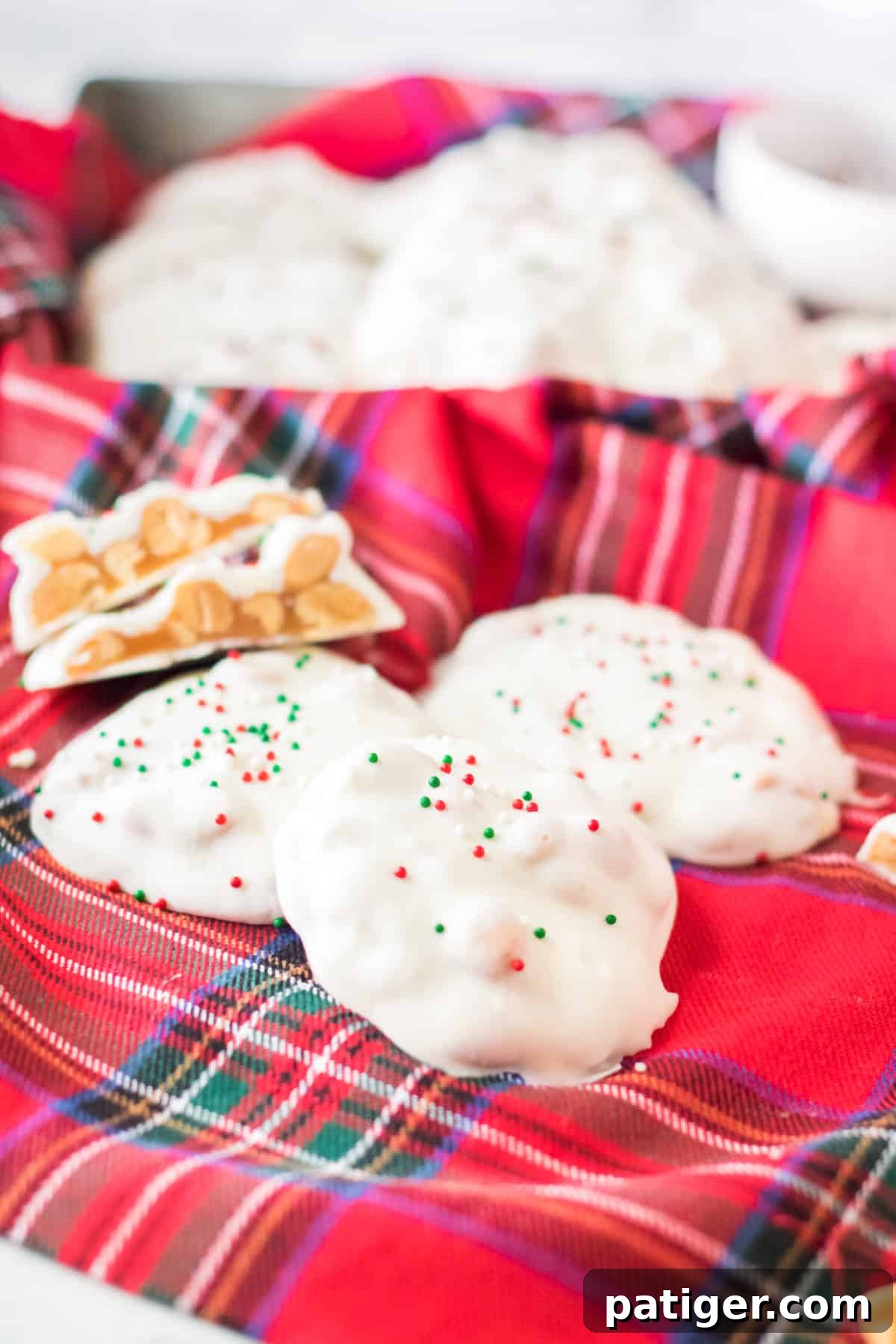 White chocolate polar bear paws candy with festive red and green sprinkles on a red plaid linen. One candy is halved, revealing its luscious caramel and peanut core.
