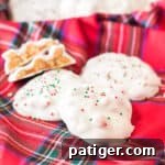White chocolate polar bear paws candy with red and green sprinkles on top on a red plaid linen. One candy is cut in half to show caramel and peanuts inside.