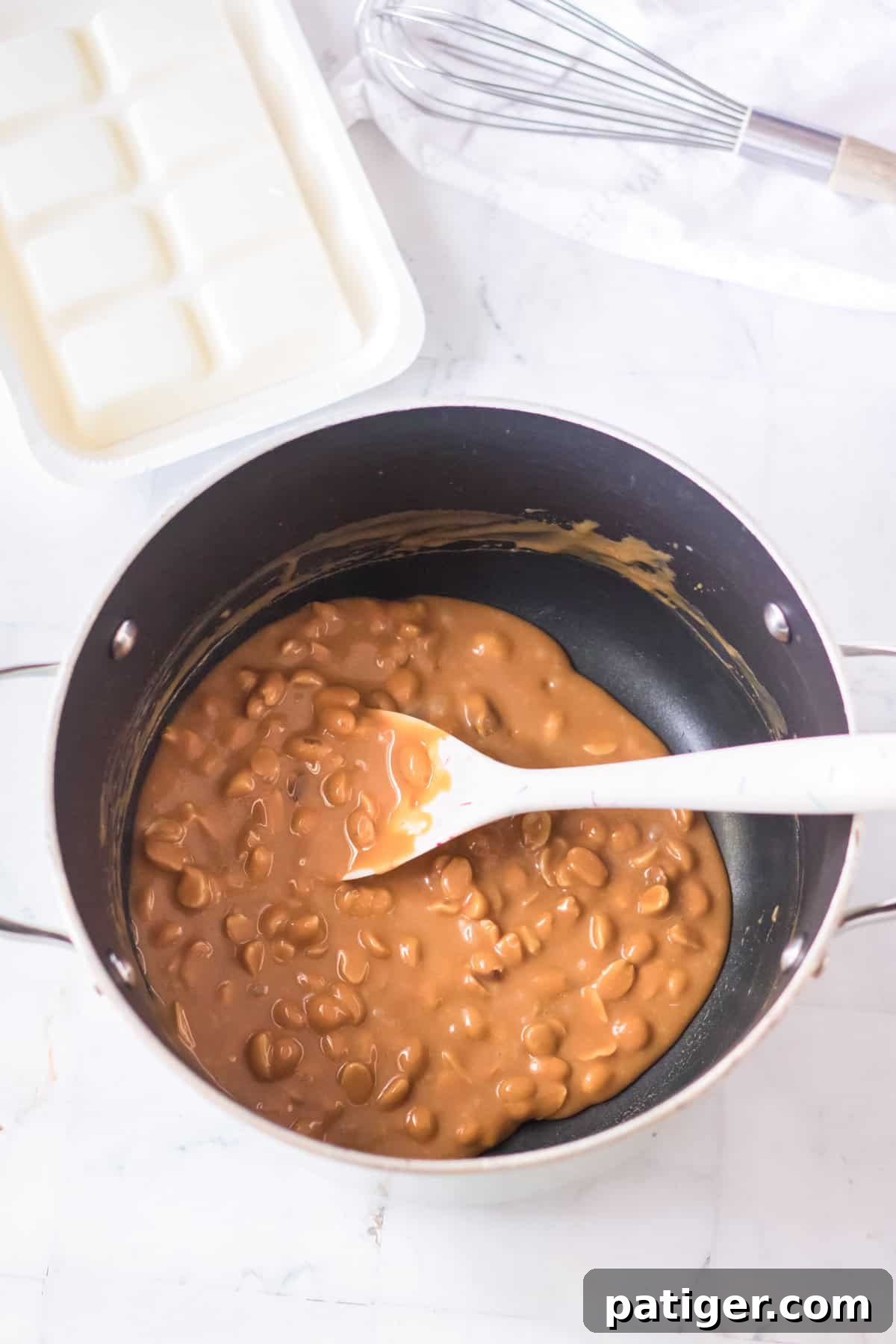 Roasted salted peanuts being stirred into the smooth caramel mixture in a saucepan with a wooden spoon.