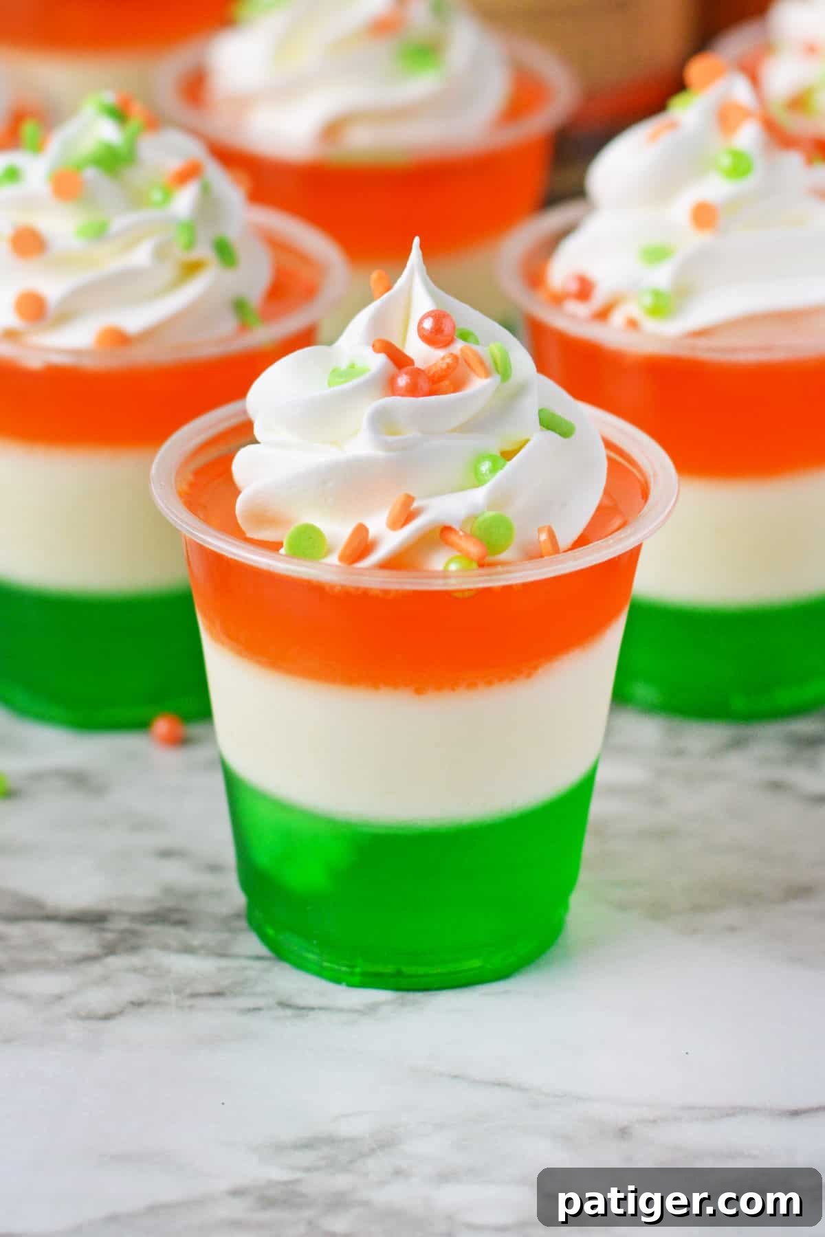 St. Patrick's Day jello shots made with a green bottom layer, white middle layer, and orange top layer, and whipped cream and festive sprinkles.