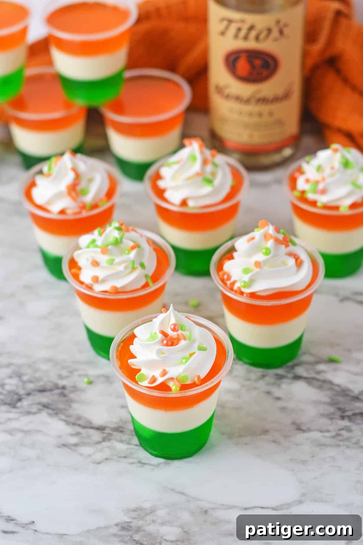 Whipped cream and sprinkle topped Irish flag jello shots with bottle of Tito's vodka behind them.