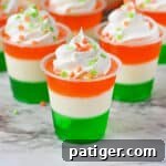 Festive St. Patrick's Day jello shots made with layers of green, white, and orange jello and topped with whipped cream and green and orange sprinkles.
