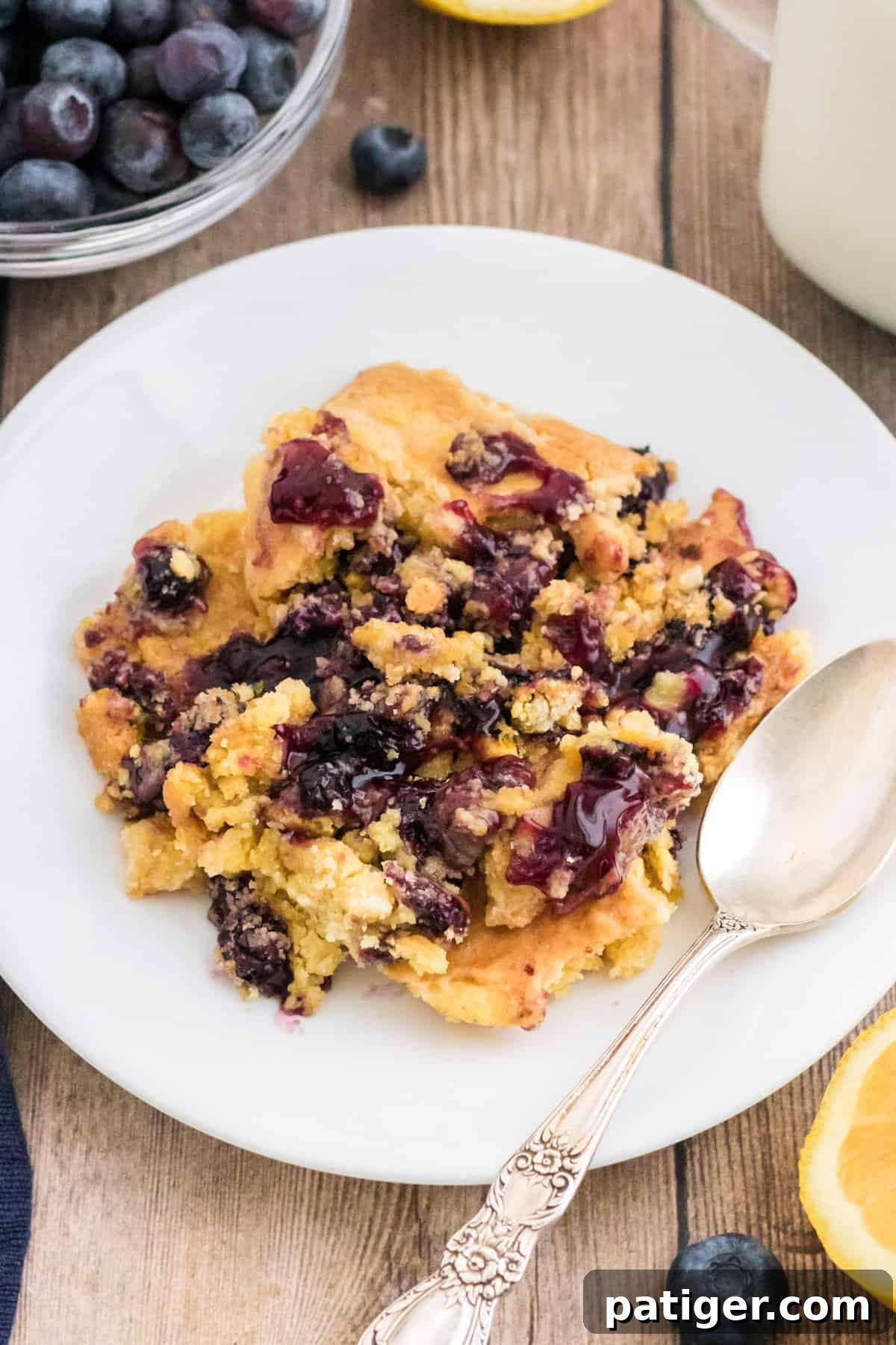 Lemon blueberry dump cake served on a white plate with a silver spoon and milk, showcasing its golden crust and vibrant fruit filling.