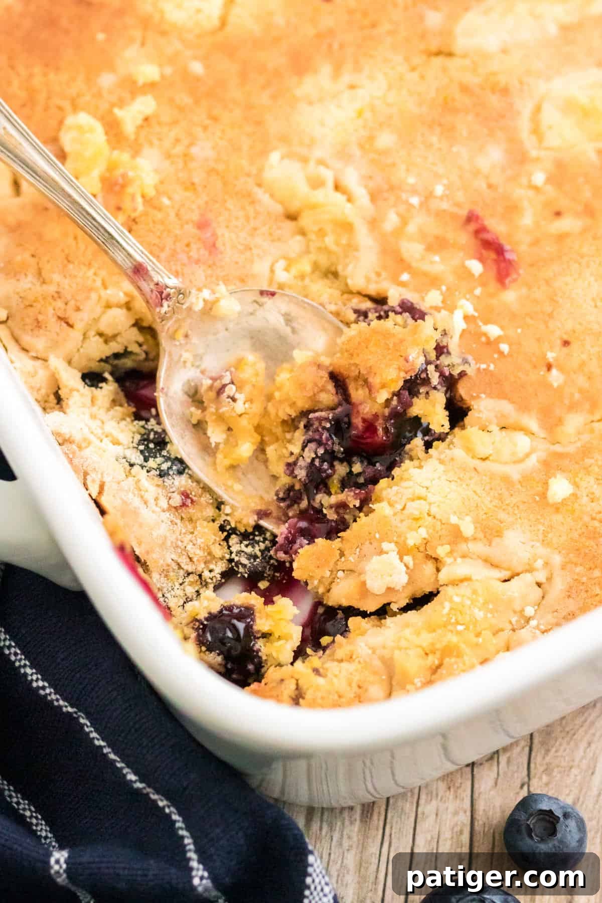 Lemon Blueberry Dump Cake baking in a white 9x13 pan, with a serving spoon gently breaking through the golden cake top to reveal the bubbling blueberry pie filling underneath.