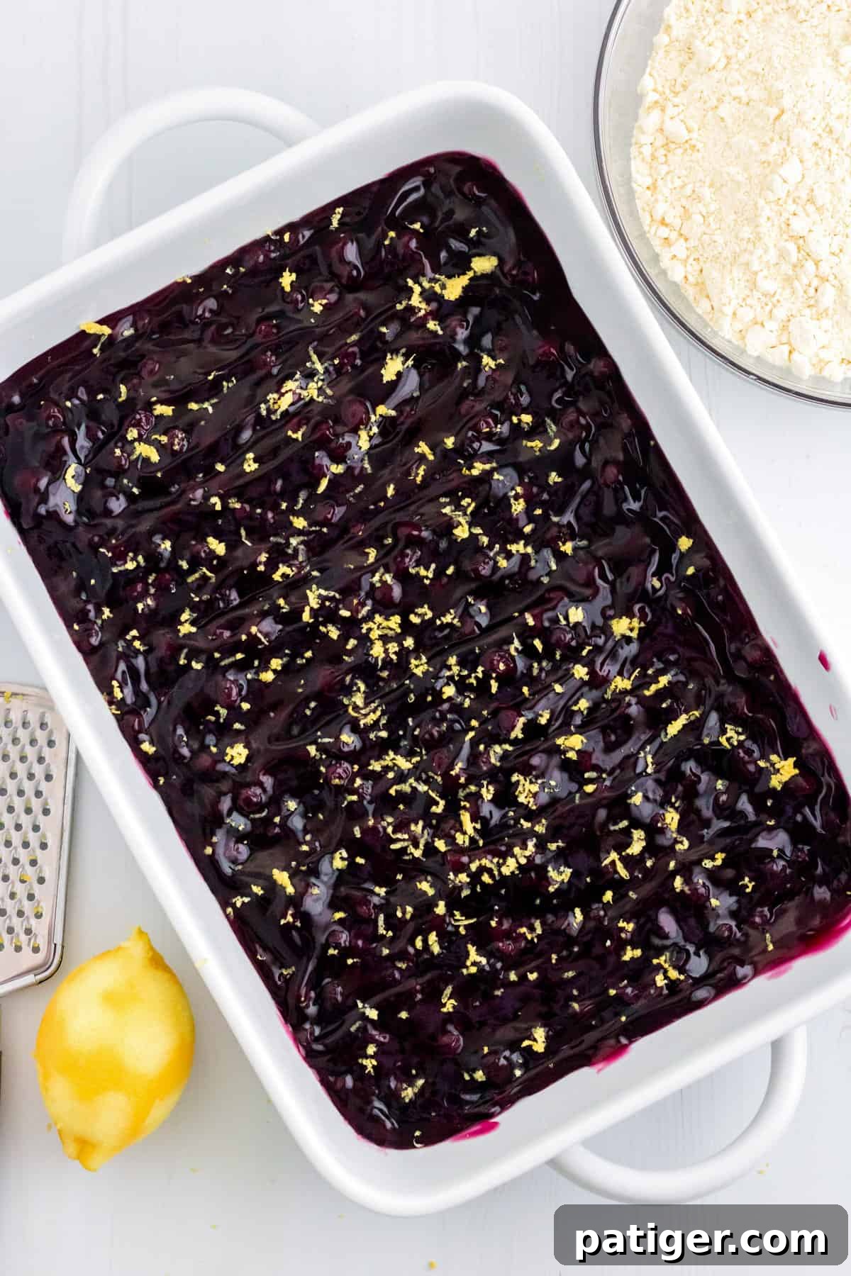 Blueberry pie filling topped with freshly grated lemon zest in a white baking dish, ready for the next layer.