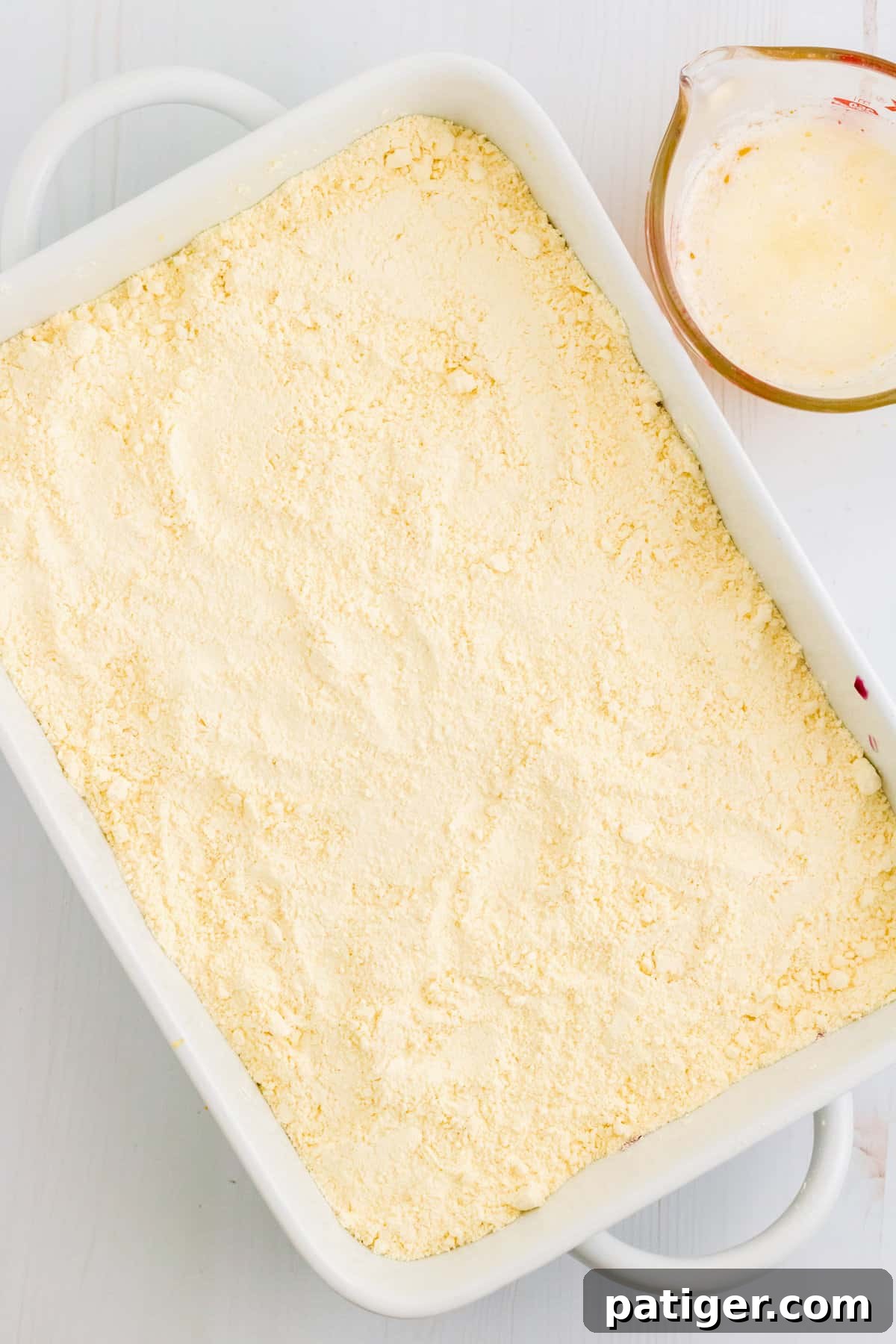 The blueberry pie filling in the baking dish has been completely covered with a layer of dry lemon cake mix, preparing it for the final butter layer.
