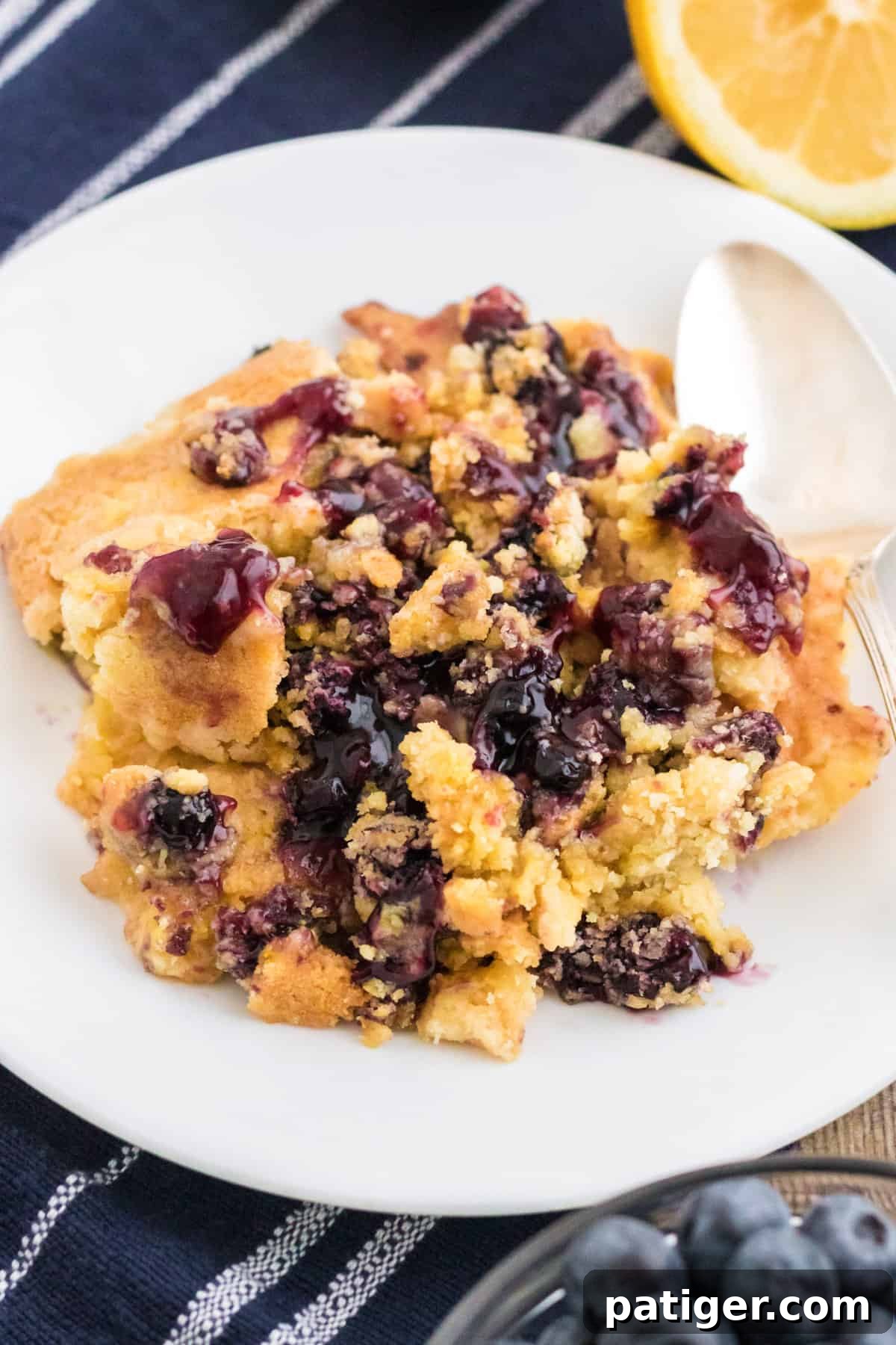 A generous serving of lemon blueberry dump cake scooped onto a pristine white plate with a silver spoon, highlighting its inviting texture.