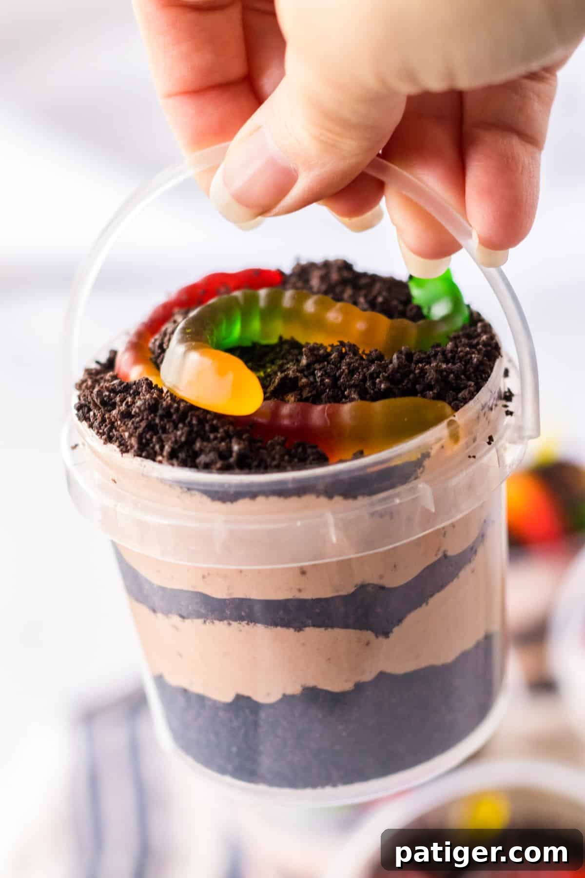Hand holding oreo dirt pudding with gummy worms in small clear plastic pail.