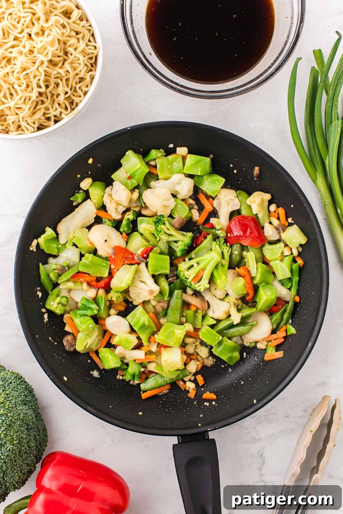 A colorful medley of stir-fry vegetables, including crisp broccoli, tender cauliflower, sweet snap peas, vibrant bell peppers, and crunchy carrots, cooking in a frying pan alongside minced garlic. Stir-fry sauce and other fresh ingredients are neatly arranged beside the pan, ready to be incorporated.