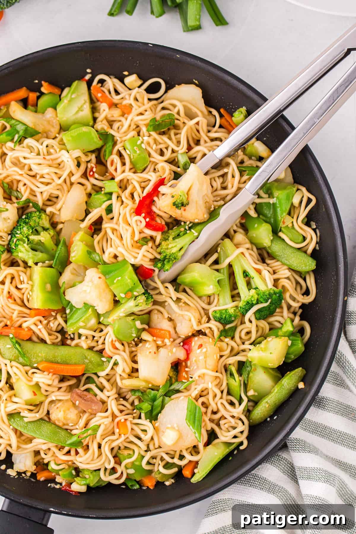 A delicious ramen noodle stir fry is presented in a black pan with a pair of serving tongs, artfully arranged and ready to be dished out and enjoyed by eager diners.