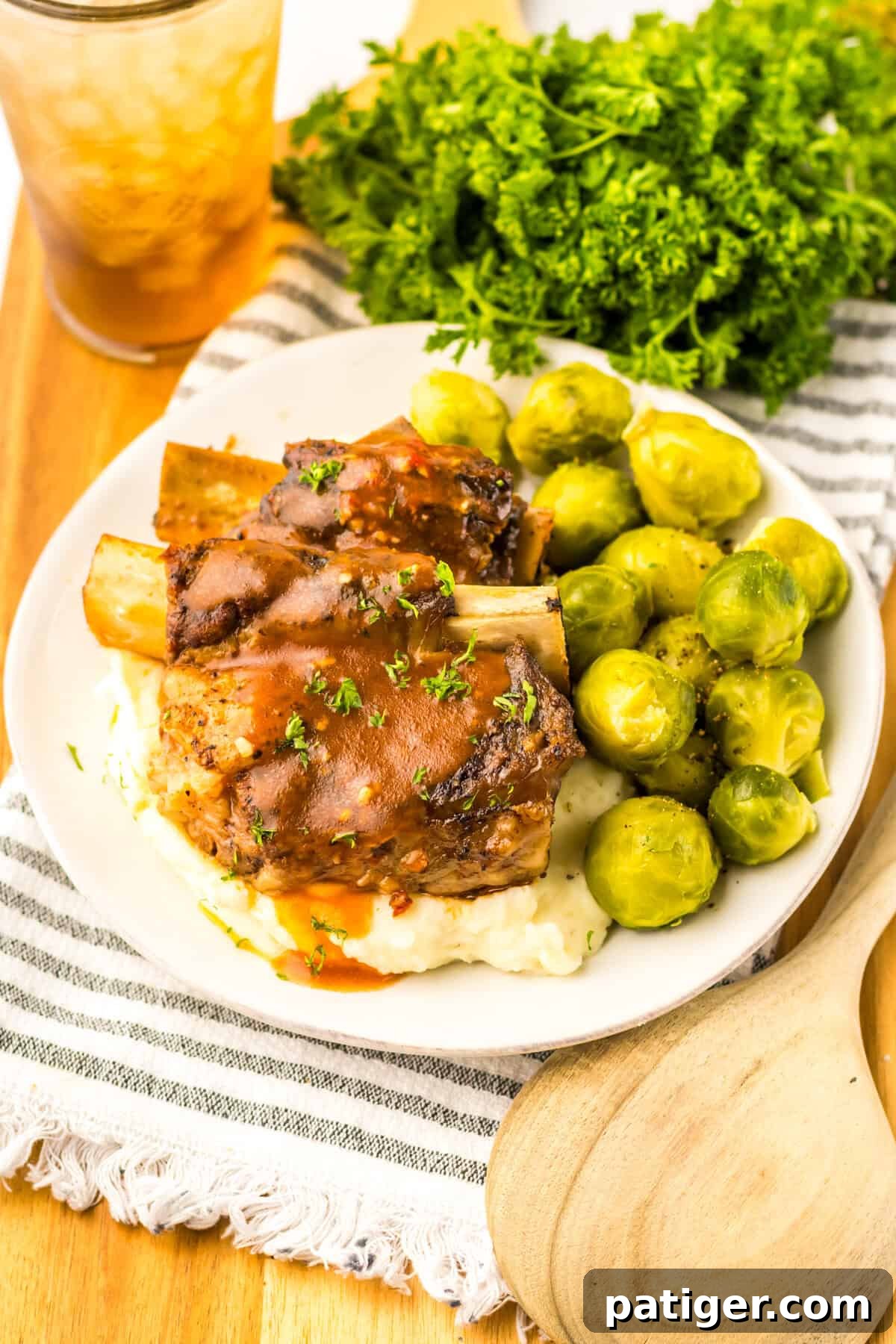 Braised beef short ribs served with creamy mashed potatoes, crisp brussel sprouts, garnished with fresh parsley, and accompanied by a glass of iced tea for a complete meal.