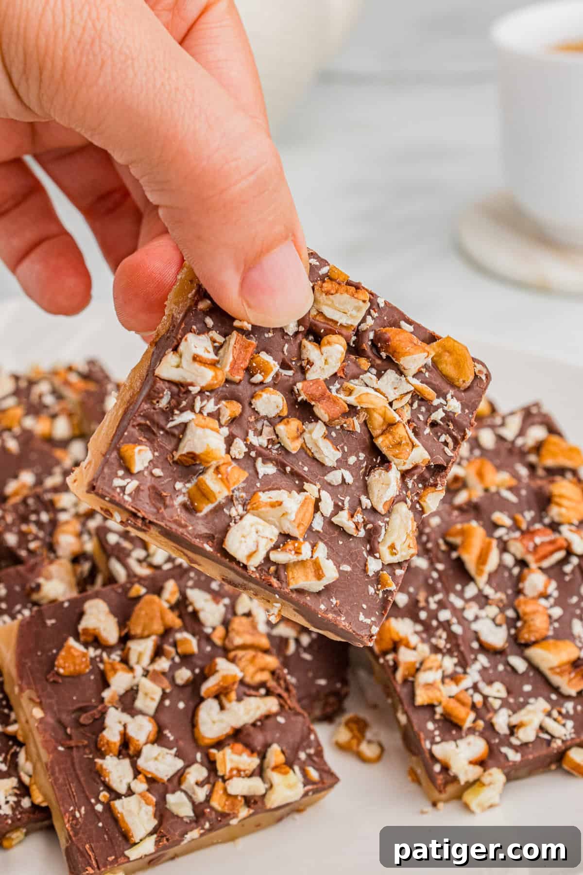 A hand holding a piece of butter toffee topped with chocolate and pecans.