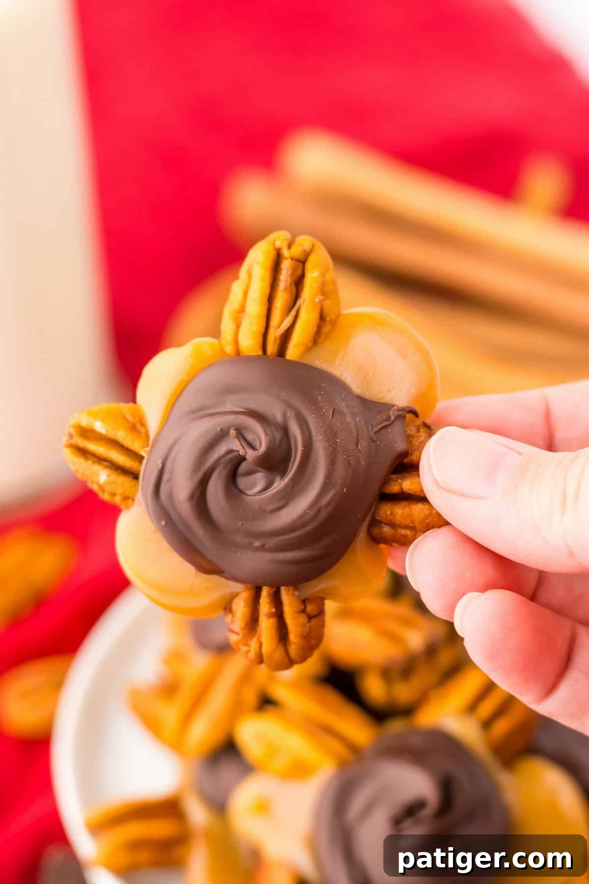Close-up of a hand holding a pecan turtle candy, featuring a cluster of pecans, gooey caramel, and a swirl of melted chocolate on top.