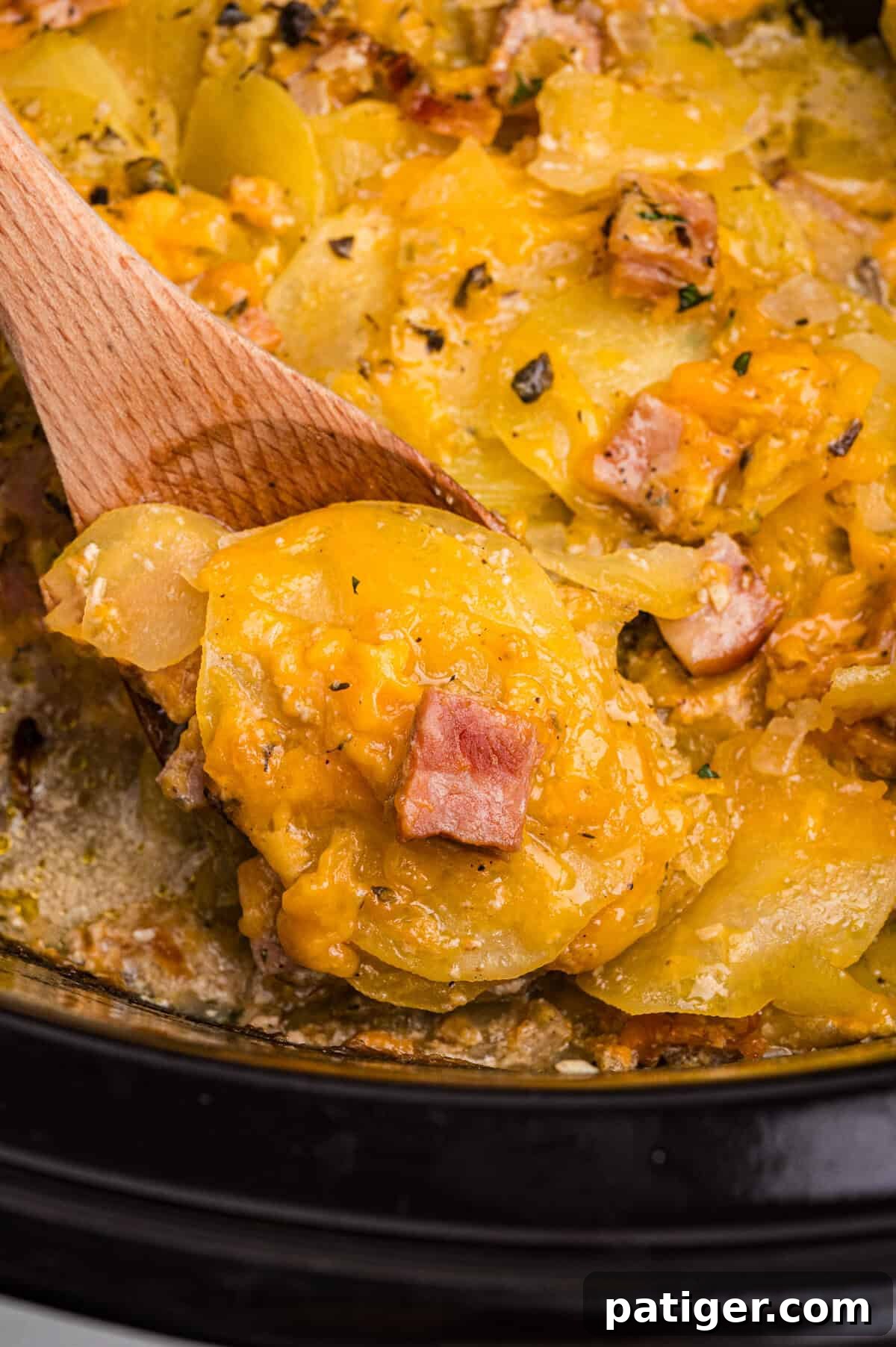 A spoon lifting a portion of cheesy scalloped potatoes and ham from a slow cooker, showing layers of tender potatoes, creamy cheese sauce, and browned ham.
