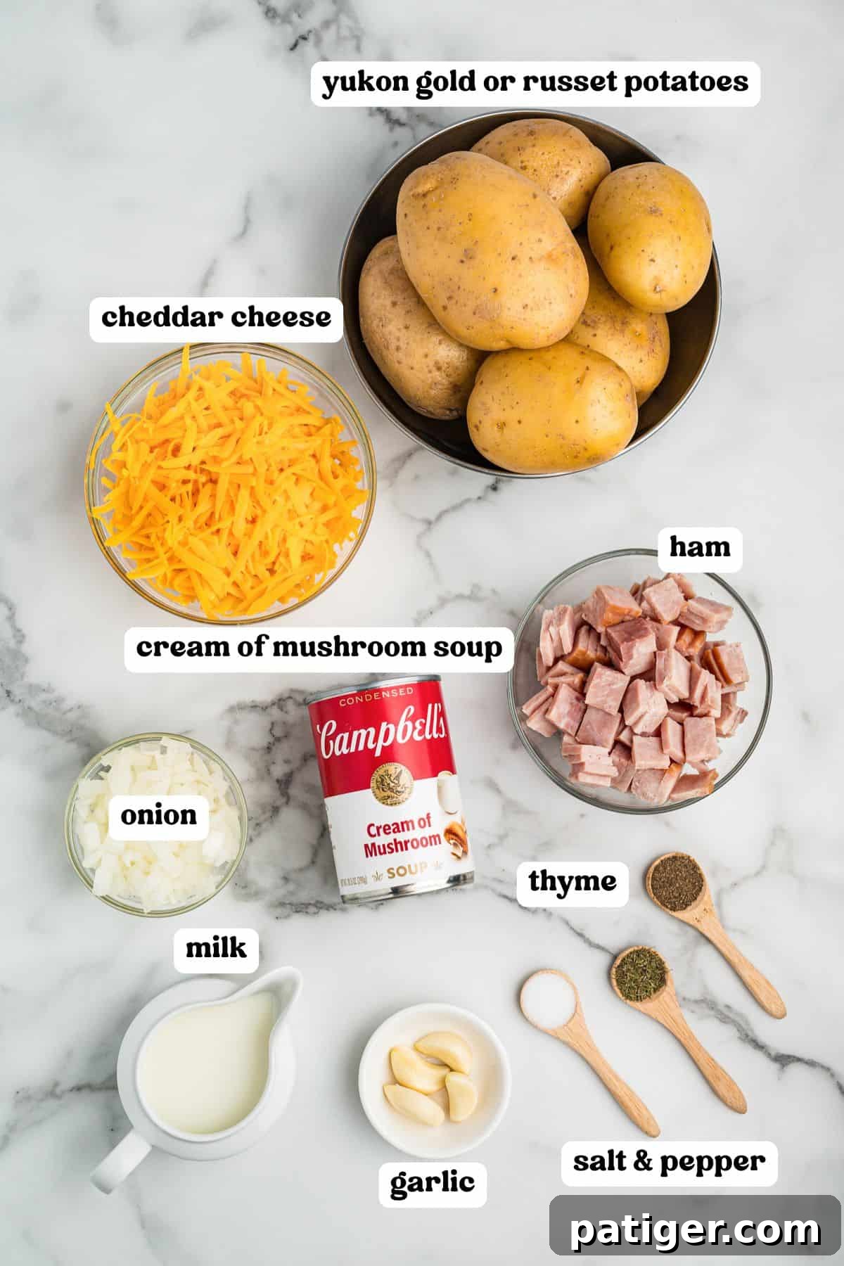 Top-down view of labeled ingredients for scalloped potatoes and ham, including Yukon Gold or Russet potatoes, shredded cheddar cheese, diced ham, a can of cream of mushroom soup, chopped onions, milk, garlic cloves, and wooden spoons filled with thyme, salt, and pepper.