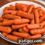 A close-up of a white bowl filled with glossy, brown sugar honey glazed baby carrots, garnished with finely chopped green herbs. The carrots have a slightly caramelized exterior and are arranged in a heaping pile.