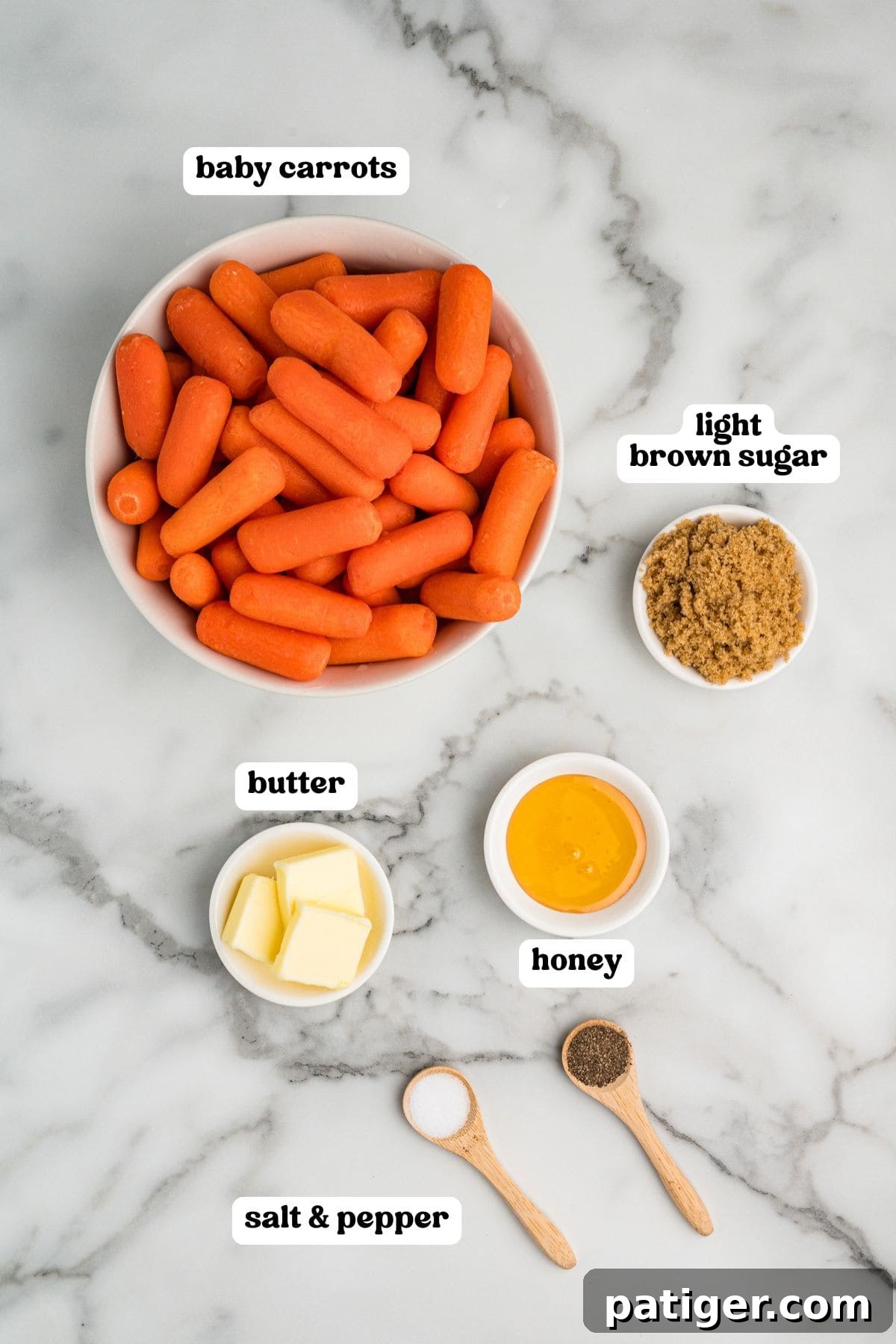 A top-down view of all the fresh ingredients laid out on a white marble surface, including a bowl of bright orange baby carrots, small dishes of light brown sugar, cubed butter, golden honey, and spoons of salt and pepper.