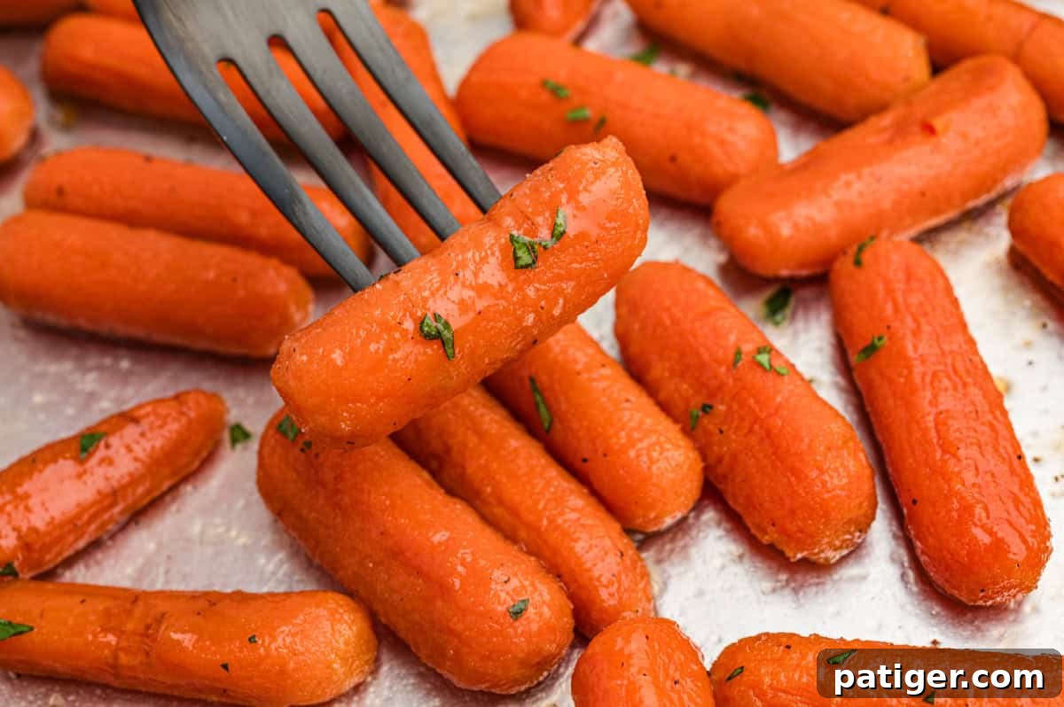 A fork gently lifting a glossy, caramelized brown sugar honey glazed carrot from a tray of perfectly roasted baby carrots, highlighting its tender texture and delicious coating.