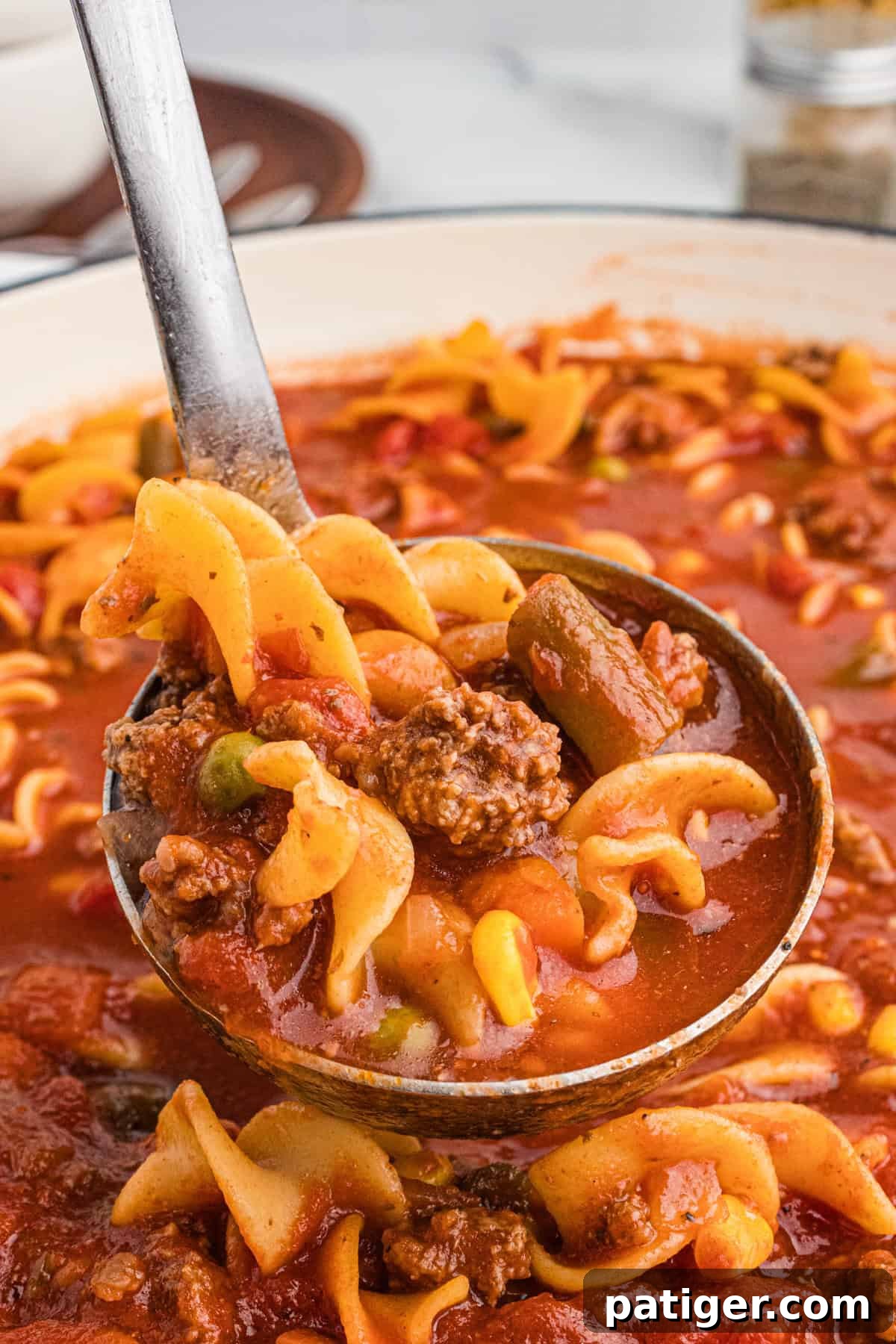 Close-up of a ladle full of rich hamburger noodle soup, perfectly showcasing the tender egg noodles, savory ground beef chunks, vibrant corn, carrots, green beans, peas, and tomatoes in a deep red broth.