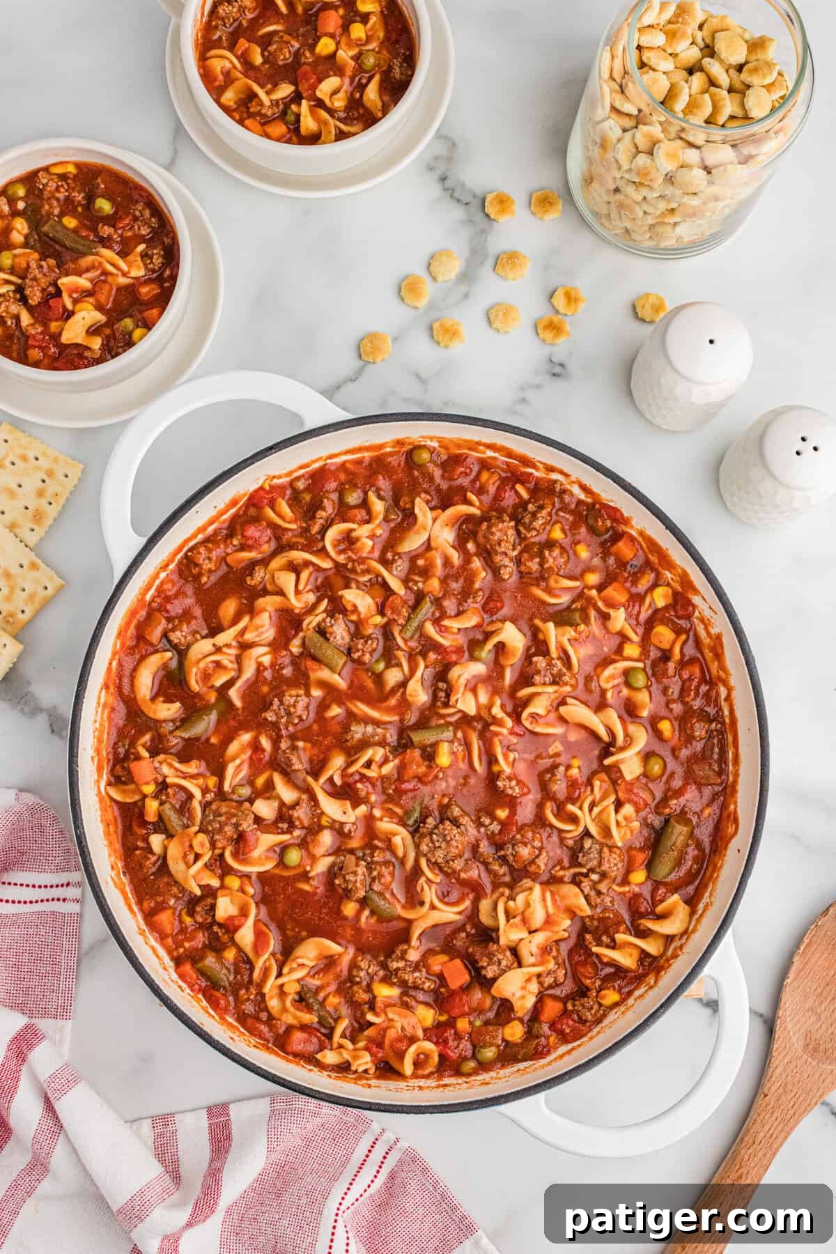 Top-down image of a white pot filled with hearty hamburger noodle soup, featuring ground beef, egg noodles, mixed vegetables, and a rich tomato-based broth. Two smaller bowls with soup are nearby, accompanied by a glass jar of oyster crackers, scattered crackers, salt and pepper shakers, and saltine crackers, ready for a comforting meal.