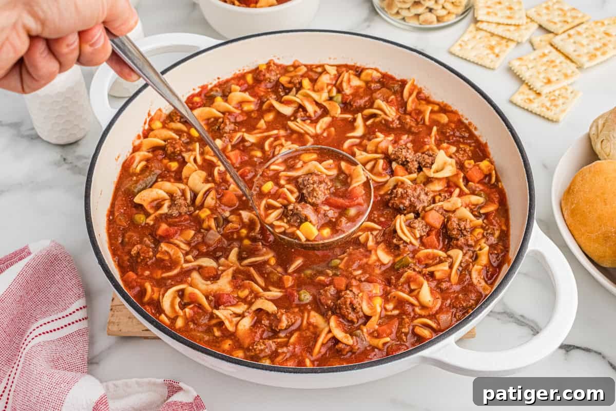 A ladle scooping steaming hamburger soup from a large pot, surrounded by various crackers and rolls. The soup is rich with ground beef, egg noodles, corn, peas, green beans, carrots, and diced tomatoes, highlighting its hearty ingredients.