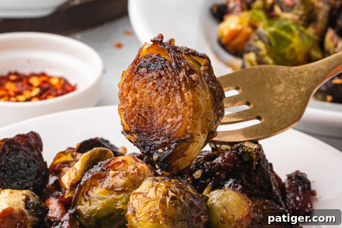 Close-up of a fork holding a single caramelized Brussels sprout with a crisp, browned surface. The background shows more Brussels sprouts on a plate and a small bowl of red pepper flakes.